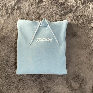 Ljusblå hoodie från Hollister XS - Supermjuk ljusblå hoodie från Hollister med vit broderad logga på bröstet och snörning i huvan. Perfekt för dig som gillar en chill och enkel stil. Hoodien är i bomullsmix och har en klassisk passform.