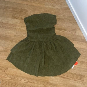 Grön axelbandslös kortklänning Cider - Supersöt grön axelbandslös kortklänning från Cider med smockad överdel och volangig kjol. Klänningen är i ett lätt, strukturerat polyestertyg och har en snygg, figurnära passform upptill som blir flowy nertill. Perfekt för sommarens alla tillfällen! Prislapp finns kvar, aldrig använd!