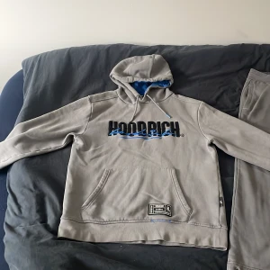 Grå dress från Hoodrich M - Säljer en fet dress från Hoodrich i storlek M. Byxorna har tryck och broderade detaljer i blått och vitt på ena benet, elastisk midja och muddar vid bensluten. Sköna och stilrena, perfekta för chill eller streetwear. Hoodien är även väldigt bekväm där de står skriver ”Hoodrich” Perfekt dress för dig som letar efter något ”chill” 