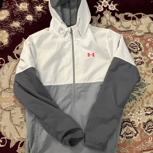 Gråvit vindjacka från Under Armour - Snygg tvåfärgad vindjacka från Under Armour i grått och vitt med huva och dragkedja framtill. Jackan har elastiska muddar och en diskret logga i rosa på bröstet. Perfekt för blåsiga dagar och sportiga outfits.