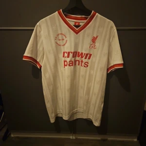 Liverpool retro fotbollströja LFC  - Äkta köpt på Unisport i Stockholm.                       Klassisk Liverpool FC fotbollströja i vitt med subtila ränder och broderat klubbmärke. Röd och gul detalj på krage och ärmslut, samt 'crown paints' tryck på bröstet. Tillverkad i 62% återvunnen polyester och 38% polyester. Perfekt för dig som älskar retrostil.