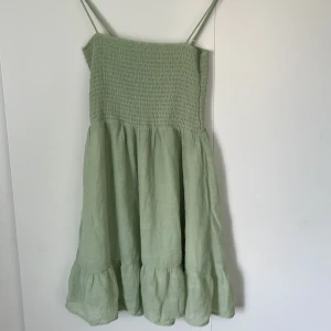 Ljusgrön sommarklänning med volang - Supersöt ljusgrön klänning med smala axelband och smock upptill. Kjolen är luftig med volangkant nertill, perfekt för varma dagar. Tillverkad i ett mjukt linneliknande material som känns svalt och skönt.