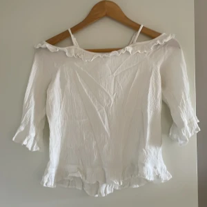 Vit offshoulder blus från Lindex - Söt vit blus från Lindex med offshoulder och volangdetaljer längs halsringning, ärmslut och nederkant. Blusen har trekvartsärm och tunna axelband för extra support. Perfekt för en fräsch och somrig look.