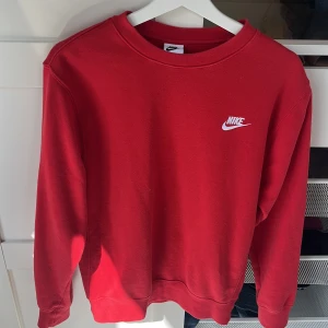 Röd Nike Hoodie - Röd sweatshirt från Nike med klassisk broderad vit logga på bröstet. Tröjan har rund halsringning, långa ärmar och är tillverkad i mjuk bomullsmix. Perfekt för en sportig och avslappnad stil.