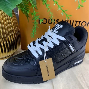Louis Vuitton svarta sneakers med logga - Svarta Louis Vuitton sneakers med vit LV-logga på plös och häl, samt präglade monogramdetaljer på sidor och tå. Skorna har vit snörning, platt sula och är tillverkade i en mix av skinn och textil. Snygg och lyxig design som sticker ut.
