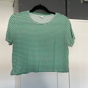 Grönvit randig T-shirt från Monki - Säljer en grön och vit randig tröja från Monki i storlek M. T-shirten har rund halsringning, korta ärmar och är gjord i mjuk bomull. Perfekt för en chill och avslappnad stil. Passformen är normal och toppen är lätt att matcha med jeans eller kjol.