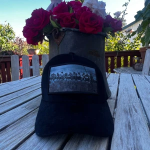 Svart keps med vintage fototryck - Cool svart keps med ett klassiskt vintage fototryck framtill som föreställer arbetare på en balk högt över New York. Kepsen har böjd skärm och justerbar baksida. Perfekt för dig som gillar streetstyle och unika accessoarer.