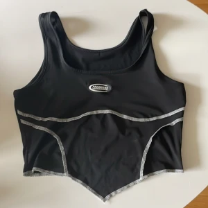 Svart sportig linnetopp Progress - Svart linnetopp från Progress med grå kontrastsömmar och sportig look. Toppen har breda axelband, croppad passform och spetsig nederkant. Perfekt för träning eller en aktiv stil. Snygg logga framtill och stretchigt syntetmaterial.