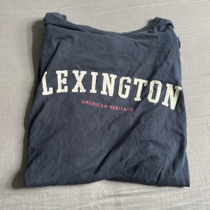 Mörkblå Lexington t-shirt med tryck - Snygg mörkblå t-shirt från Lexington med stort vitt tryck över bröstet och texten 'AMERICAN HERITAGE' i rött. Klassisk passform och rund halsringning, perfekt till jeans eller shorts. Gjord i mjuk bomull för skön känsla hela dagen. Hör gärna av er vid minst lilla fråga 