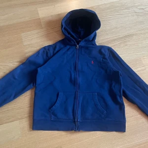 Marinblå hoodie från Polo Ralph Lauren - Marinblå hoodie från Polo Ralph Lauren med dragkedja och huva. Klassisk röd broderad logga på bröstet och känguruficka framtill. Tillverkad i mjuk bomullsblandning med polyester. Perfekt för chill dagar eller när du vill ha en clean look.