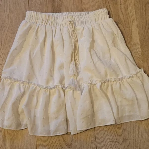 Vit volangkjol med knytband - Supersöt vit kortkjol med volangkant och diskret strukturmönster. Kjolen har knytband med tofsar framtill. Perfekt för sommarens alla tillfällen.