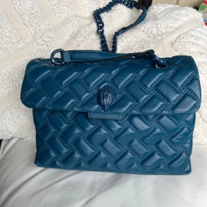 Blå quiltad axelväska från Kurt Geiger - Snygg blå axelväska från Kurt Geiger i äkta skinn med quiltad framsida och kedjerem. Väskan har lock med dekorativ detalj och rymligt innerfack med logomönstrat foder. Perfekt för dig som vill ha en stilren men ändå unik väska.