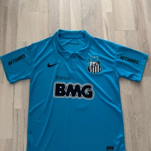 Santos Neymar Jr blå fotbollströja Nike - Blå fotbollströja från Nike med Santos FC-emblem, Neymar Jr och nummer 11 på ryggen. Tröjan har korta ärmar, krage med knappar och tryckta sponsorer som BMG och Netshoes. Perfekt för dig som älskar brasiliansk fotboll och Neymar.