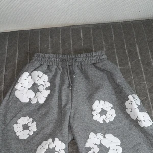 Grå Denim Tears shorts  - Grå mjukisbyxor från Dem Tears med elastisk midja och snörning. Byxorna har stora vita tryck i form av blommor eller cirklar över hela plagget. Tillverkade i mjuk bomull, perfekt för en chill och avslappnad stil. Priset kan diskuteras 