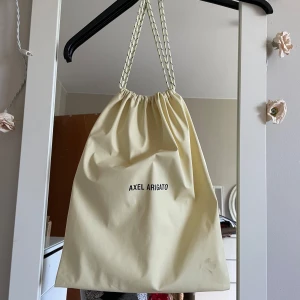 Beige Axel Arigato ryggsäck med snörning - Säljer en stilren beige ryggsäck från Axel Arigato med snörning upptill och dubbla dragsko-band. Väskan är tillverkad i ett mjukt syntetmaterial och har minimalistisk design med svart logga framtill. Perfekt för att bära med sig det viktigaste till skolan eller stan.