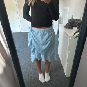 Ljusblå omlottkjol med volang - Supersöt ljusblå omlottkjol med små vita prickar och volangkant. Kjolen knyts i midjan och har en flowy passform som känns riktigt somrig och fräsch. Perfekt för dig som gillar en feminin och avslappnad stil.