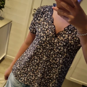 Blommig blus från ONLY XS - Supersöt blommig blus från ONLY i marinblått med små vita och beige blommor. Blusen har korta ärmar, vågig v-ringad kant och luftig passform. Perfekt till jeans eller kjol för en fräsch look.