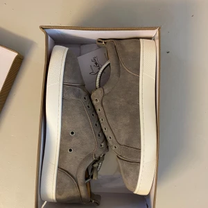 Skitsnygga Christian Louboutin skor - Snygga grå sneakers från Christian Louboutin i mjuk mocka med klassisk vit sula och ikonisk röd undersida. Skorna har rund tå, snörning och diskreta metalldetaljer. Perfekta för dig som vill sticka ut med exklusiv stil. Hör av dig vid funderingar.