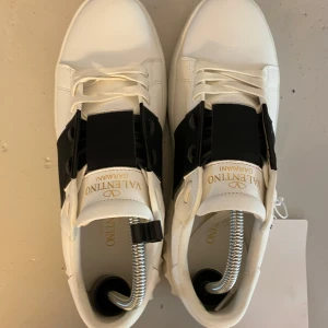 Valentino opens - Snygga vita Valentino Garavani One Stud XL sneakers med bred svart rem över vristen och coola nitar på sulan. Skorna har rund tå, snörning och är tillverkade i skinn. Perfekta för dig som gillar exklusiv streetstyle och vill sticka ut med en lyxig touch.