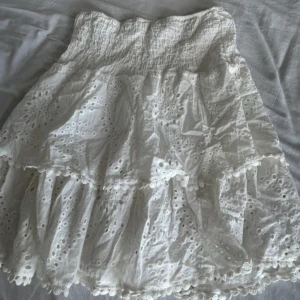 Vit volangkjol med broderade detaljer - Supersöt vit kortkjol med volanger och broderade hålmönster. Kjolen har resår i midjan och små bollar längs kanterna för extra detalj. Jättesöt men har aldrig fått andvändning för den. Köptes för 280.