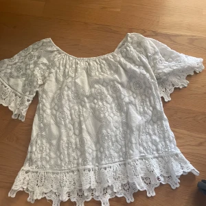 Vit blus med spets och blommönster - Superfin vit blus med breda ärmar och vackra spetsdetaljer. Blusen har blommigt broderat mönster och volangkant nertill och vid ärmslut. Perfekt för en somrig look med boho-vibbar.