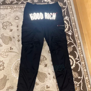 Svarta cargopants från Hoodrich - Svarta cargopants från Hoodrich med stora reflekterande HOOD RICH-tryck framtill och broderad logga på benfickan. Byxorna har flera fickor med dragkedja, elastisk midja med snörning och en loose passform. Perfekta för en streetwear-look.