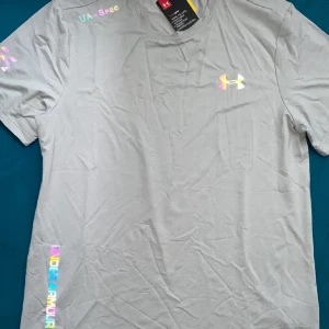 Under Armour t-shirt och shorts - Snygg grå t-shirt från Under Armour med holografiska loggor och tryckdetaljer på bröstet, axeln och nedtill. Tillverkad i mjuk och lätt polyester, perfekt för träning. Klassisk rund hals och korta ärmar för en sportig vibe. Och pris kan diskuteras 