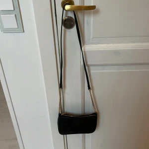 Svart axelväska från H&M med kedja - Snygg svart axelväska från H&M med guldfärgad kedjedetalj och dragkedja upptill. Väskan har en minimalistisk och stilren design med lång axelrem i syntetmaterial. Perfekt storlek för det viktigaste och passar till många outfits.