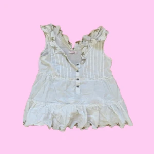 Vit volangblus med broderi från Only - Supersöt vit blus från Only med volangkant och broderade detaljer i beige längs halsringning och nederkant. Ärmlös modell med knappar framtill och lätt rynkad passform. Perfekt för en somrig look med romantiska vibes.