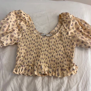 Blommig croppad blus från NA-KD - Supersöt blommig blus från NA-KD i ljusgul nyans med lila och rosa småblommor. Modellen är croppad med smockad kropp, volangkant nertill och puffiga korta ärmar. Perfekt för vår och sommar, toppen är helt oanvänd men har inte prislappen kvar🫶🏼