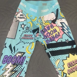 Stronger leggings med pop art-mönster - Färgglada leggings från Stronger med pop art-inspirerat tryck, text som 'Girl Power' och 'Boom!', samt bananer och blixtar. Basen är ljusblå med inslag av gult, rosa, svart och lila. Hög midja och tight passform, perfekta för träning eller när du vill sticka ut.