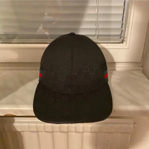 Svart Gucci keps med GG-mönster - Snygg svart keps från Gucci med diskret GG-monogram över hela kepsen. Klassisk böjd skärm och justerbart spänne bak i svart skinn. Sidorna har gröna och röda ränder som ger den ikoniska Gucci-looken. Perfekt accessoar för att lyfta din outfit.