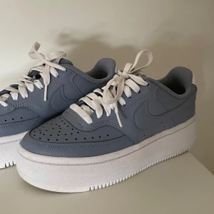 Nike Air Force 1 i ljusblått - Snygga Nike Air Force 1 sneakers i en fräsch ljusblå färg med vita snören och tjock vit plattformssula. Skorna har klassisk rund tå och är tillverkade i syntetmaterial med perforerade detaljer framtill för extra stilpoäng.