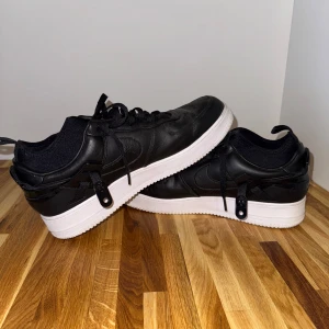 Svarta Nike Air Force 1 Undercover black sneakers - Snygga svarta Nike Air Force 1 sneakers i skinn med vit platt sula. Klassisk låg modell med snörning och diskreta detaljer som dragkedja på sidan. Perfekta för dig som gillar streetstyle och vill ha en clean look.