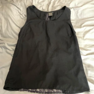 Svart linnetopp med spets från ICHI - Svart ärmlös linnetopp från ICHI i storlek M. Toppen har en snygg spetsdetalj i ryggen som ger en cool kontrast och extra edge. Rund halsringning och rak passform, perfekt att styla med jeans eller kjol för en trendig look.