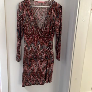 Mönstrad omlottklänning Pernilla Wahlgren - Färgstark knälång omlottklänning från Pernilla Wahlgren med lång ärm och djup v-ringning. Klänningen är i stretchigt material har ett coolt geometriskt mönster i rött, svart, beige, blått och vitt. Sitter snyggt med figurnära passform och är perfekt för dig som gillar att sticka ut. Snygg till tajta jeans. 