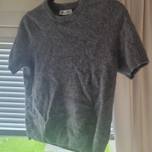 Grå stickad tröja från Zara - Mysig grå stickad tröja från Zara med korta ärmar och rund halsringning. Tröjan har en enkel och stilren design med ribbade muddar vid ärmslut och nederkant. Perfekt för lager-på-lager och chill vardagsstil.