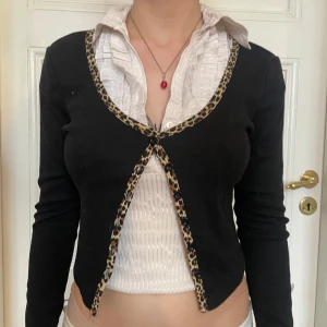 Svart kofta med leopardkant - Säljer en svart croppad kofta med lång ärm och snygga leopardmönstrade kanter. Koftan har knappar framtill så du kan styla den öppen eller stängd. Perfekt för dig som vill ha en edgy och trendig look med lite Y2K-vibe.