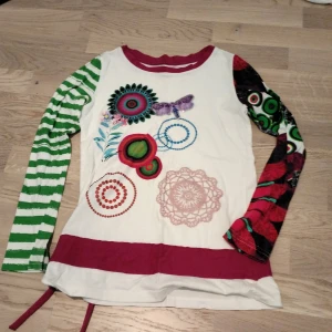 Desigual långärmad mönstrad tröja - Färgstark långärmad tröja från Desigual med unika mönster och tryck framtill. Ena ärmen är grön- och vitrandig, den andra har färgglada cirklar och rött mönster. Tröjan har vinröda detaljer vid hals och nederkant samt knytband i sidan. Perfekt för dig som gillar att sticka ut.