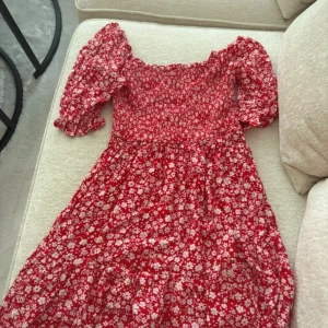 Röd blommig klänning med puffärm - Superfin röd klänning med vita blommor och puffiga trekvartsärmar. Smockad överdel och volang i kjolen ger en söt och trendig look. Perfekt för dig som gillar färg och mönster! Klänningen har en feminin passform och är lätt att bära.