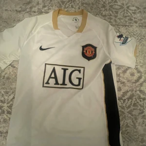 Manchester United Ronaldo 7 vit Nike tröja - Säljer en klassisk Manchester United fotbollströja med Ronaldo 7 på ryggen. Tröjan är vit med svarta detaljer och guldkrage, tillverkad i lätt och ventilerande material. AIG-logga på bröstet, klubbmärke och Premier League-patch på ärmen. Perfekt för fans!
