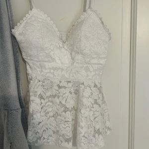 Vit spetsig blommig linnetopp - Supersöt vit linnetopp med blommig spets och tunna axelband. Toppen har vadderade kupor och fina spetsdetaljer längs kanterna. Den har en lätt utsvängd form nedtill och är perfekt för en somrig look. Passar dig som gillar Sthlm stil och feminin stil.