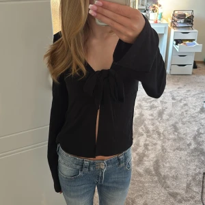 Svart knytblus från Gina Tricot - Supersnygg svart blus från Gina Tricot med lång ärm och djup v-ringning. Toppen har en stor knytrosett framtill och är croppad i modellen. 