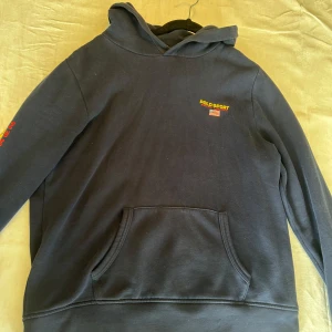 Mörkblå hoodie Polo Sport Ralph Lauren - Mörkblå hoodie från Polo Sport Ralph Lauren i storlek junior XL=ca S. Klassisk modell med huva, magficka och broderad logga samt amerikansk flagga på bröstet. Stor gul och röd logga på ryggen. Mjuk bomullskänsla och långärmad.
