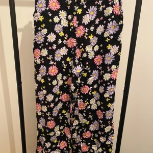 Blommiga vida byxor från H&M Divided - Säljer ett par svarta byxor från H&M Divided med färgglatt blommönster i lila, rosa, vitt och gult. Byxorna har en loose och wide passform med resår i midjan för extra komfort. Perfekta för dig som vill sticka ut med en färgglad look.