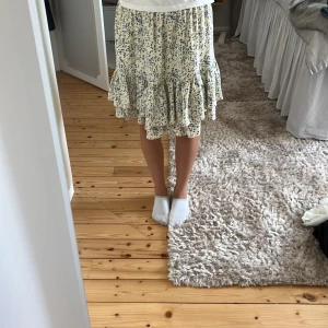 Blommig volangkjol i ljusblått - Säljer en söt kort kjol med volangkant och blommigt mönster i ljusblått och vitt. Kjolen har en avslappnad passform och är perfekt för varma dagar. Materialet känns lätt och luftigt, och volangen ger en extra touch av stil.