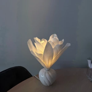 Unik stilren Blomlampa - Bilderna visar en dekorativ bordslampa med blomformad skärm i vitt papper och en rund, vit keramisk fot. Lampan ger ett mjukt, varmt sken och passar perfekt som mysig inredningsdetalj i moderna hem. Ingen bok syns på bilderna.