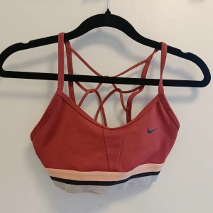 Roströd sportig linnetopp från Nike - Snygg roströd linnetopp från Nike med tunna axelband och coolt korsad rygg. Toppen har meshdetalj framtill och bred resår i beige, svart och silver nedtill. Perfekt för träning eller när du vill ha en sportig look.