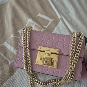 Ljusrosa Gucci axelväska i skinn - Säljer en ljusrosa axelväska från Gucci i präglat skinn med GG-logga över hela väskan. Väskan har en  guldfärgad kedja och ett snyggt lås framtill. Insidan är fodrad i beige och har ett litet innerfack. Perfekt för dig som vill sticka ut med lyxig vibe. Nypris 25000 kr Säljer för 10000 kr 