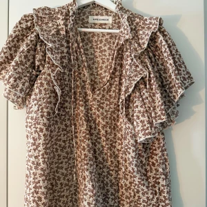 Brun och vit mönstrad blus Sofie Schnoor - Supersöt blus från Sofie Schnoor i brun och vit med ett unikt mönster. Blusen har volangdetaljer vid axlar och hals, v-ringad med knyt i halsen. Tillverkad i mjuk bomull.